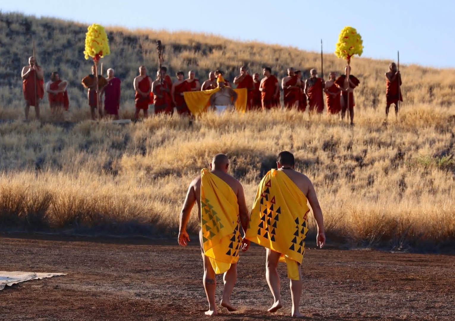 Practitioners in yellow kīhei walk toward a gathered procession in red kīhei holding kāhili at Puʻukoholā Heiau during golden hour.