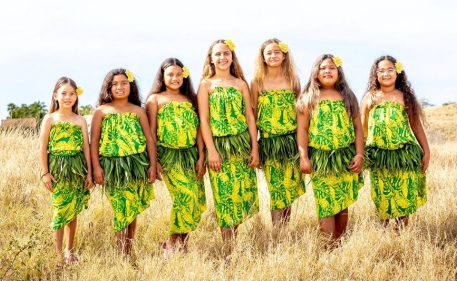 Tavana Rahi hālau keiki in matching yellow-and-green pāʻū tops and lāʻī skirts standing together in a sunlit grass field.