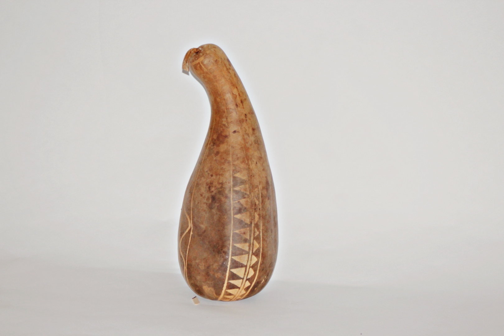 A finished ipu pāwehe gourd — a tall curved bottle gourd with carved triangular and linear patterns scored into the natural brown skin.