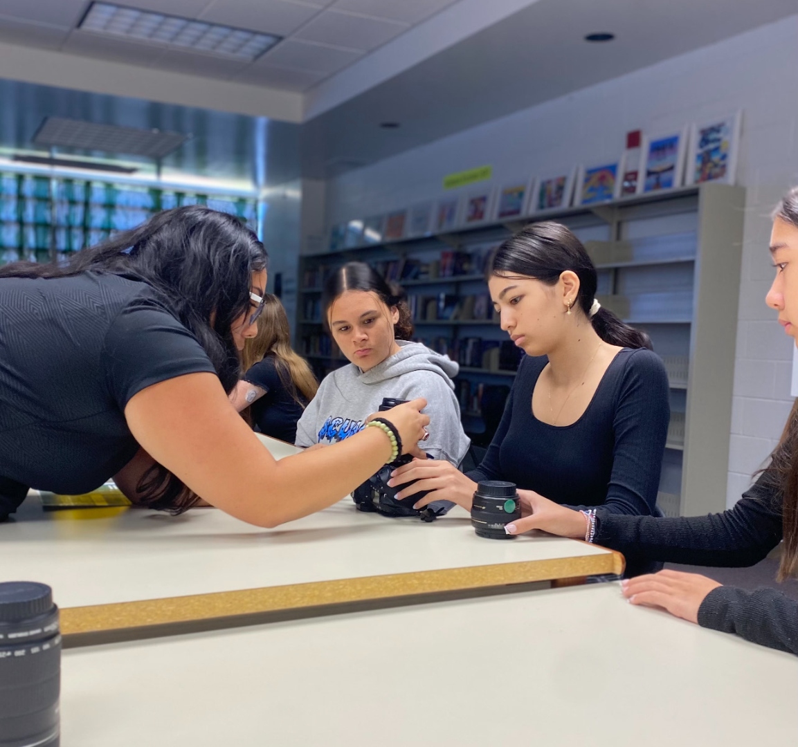 A teacher demonstrates a camera and lens to attentive intermediate-school students in a school library — Kealakehe Media Program in session.