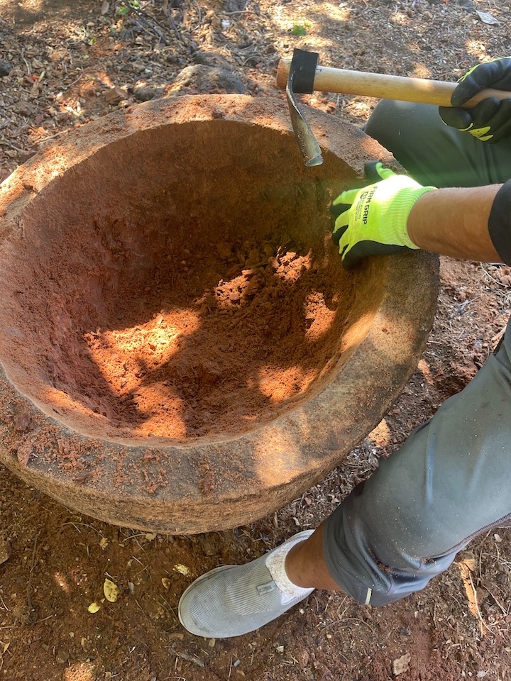 Gloved hands using a hammer and chisel to hollow out a large wooden bowl in the dirt outdoors during a Hale Mua workshop.