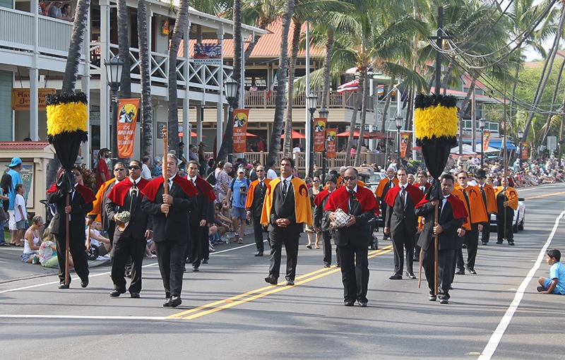 Kāhili bearers and Royal Order procession in red and orange capes carrying tall feathered standards down Aliʻi Drive during the King Kamehameha Day Celebration Parade in Kona